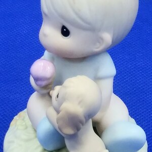以下が含まれることがあります： アイスクリームコーンと子犬を持った少年の磁器製フィギュア。少年は青いシャツと青いズボンを着用しています。犬は白く、茶色の耳をしています。フィギュアは黄色の花が咲いた緑色の台座の上に座っています。