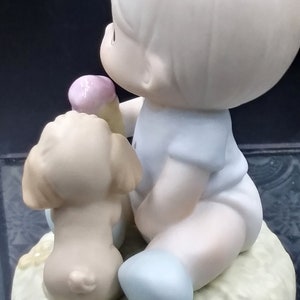 以下が含まれることがあります： 薄い青色のシャツと青い靴を履いた若い男の子の磁器製フィギュアが、小さな茶色の犬と一緒に座っています。男の子はピンクと白のアイスクリームコーンを持っています。フィギュアは薄い緑色の土台の上に座っています。