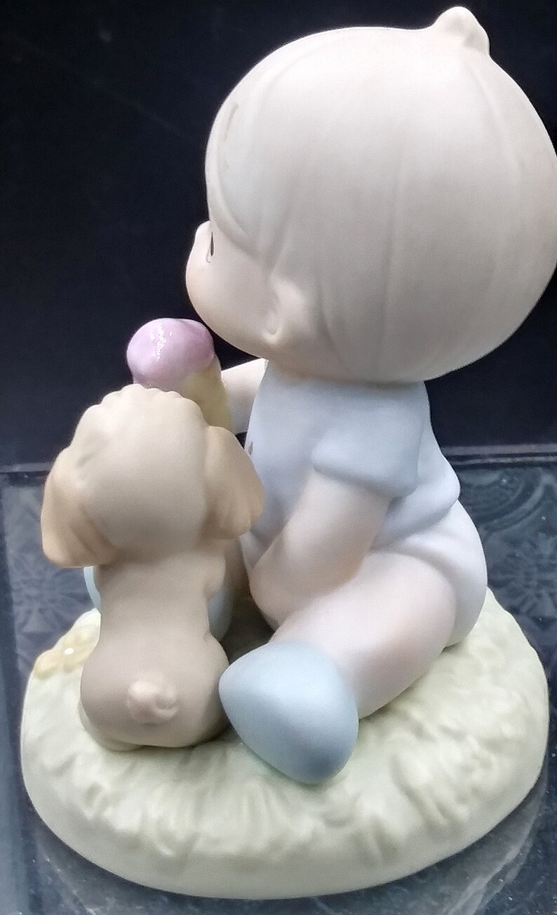 以下が含まれることがあります： 薄い青色のシャツと青い靴を履いた若い男の子の磁器製フィギュアが、小さな茶色の犬と一緒に座っています。男の子はピンクと白のアイスクリームコーンを持っています。フィギュアは薄い緑色の土台の上に座っています。