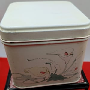 May include: A rectangular, cream-coloured metal tin with a matching lid. The tin features a floral design of white calla lilies with red accents and green leaves. The edges of the tin are trimmed with a thin pink line.