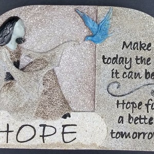 Può includere: Una placca di pietra con un'immagine scolpita di una donna che tiene un uccello. La placca ha la parola "HOPE" in grandi lettere e il testo "Make today the best it can be... Hope for a better tomorrow."