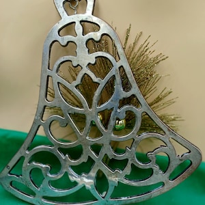 May include: A silver-colored, openwork bell-shaped ornament with intricate scrollwork. The ornament is displayed against a backdrop of gold-colored decorative sprigs and a green surface, suggesting a holiday theme.