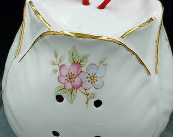 May include: White ceramic flower-shaped pot with a red ribbon, gold trim, and a floral design. The pot has holes in the bottom for drainage.