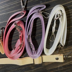 May include: A collection of colorful purse straps in pink, lavender, and white, with silver-tone metal clasps. A beige strap is also visible. The straps are made of a woven material and are designed for attaching to a purse or bag.