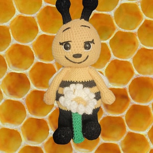 May include: A handmade crochet bee doll with a smiling face, black antennae, and black and yellow striped body. The bee doll has a white and yellow flower design on its abdomen and black legs. The background is a honeycomb pattern.