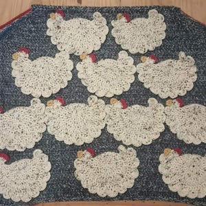 Puede incluir: Un cojín de asiento rectangular de ganchillo moteado en azul y blanco con diez aplicaciones de gallinas de ganchillo blanco con crestas rojas y picos amarillos. El cojín tiene lazos rojos en las esquinas superiores para sujetarlo a una silla.