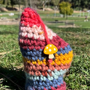 May include: A colorful, hand-crocheted cozy, featuring a mushroom charm. The cozy is made with various yarn colors, including red, pink, blue, and yellow. The mushroom charm is yellow with white spots. The cozy is designed to fit a cup or bottle.