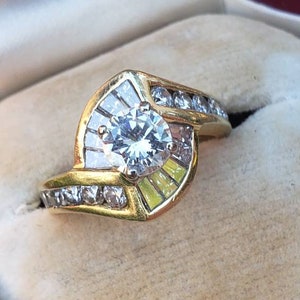 May include: A gold ring with a large, round, clear center stone surrounded by smaller clear stones. The ring is set in a curved, prong setting.