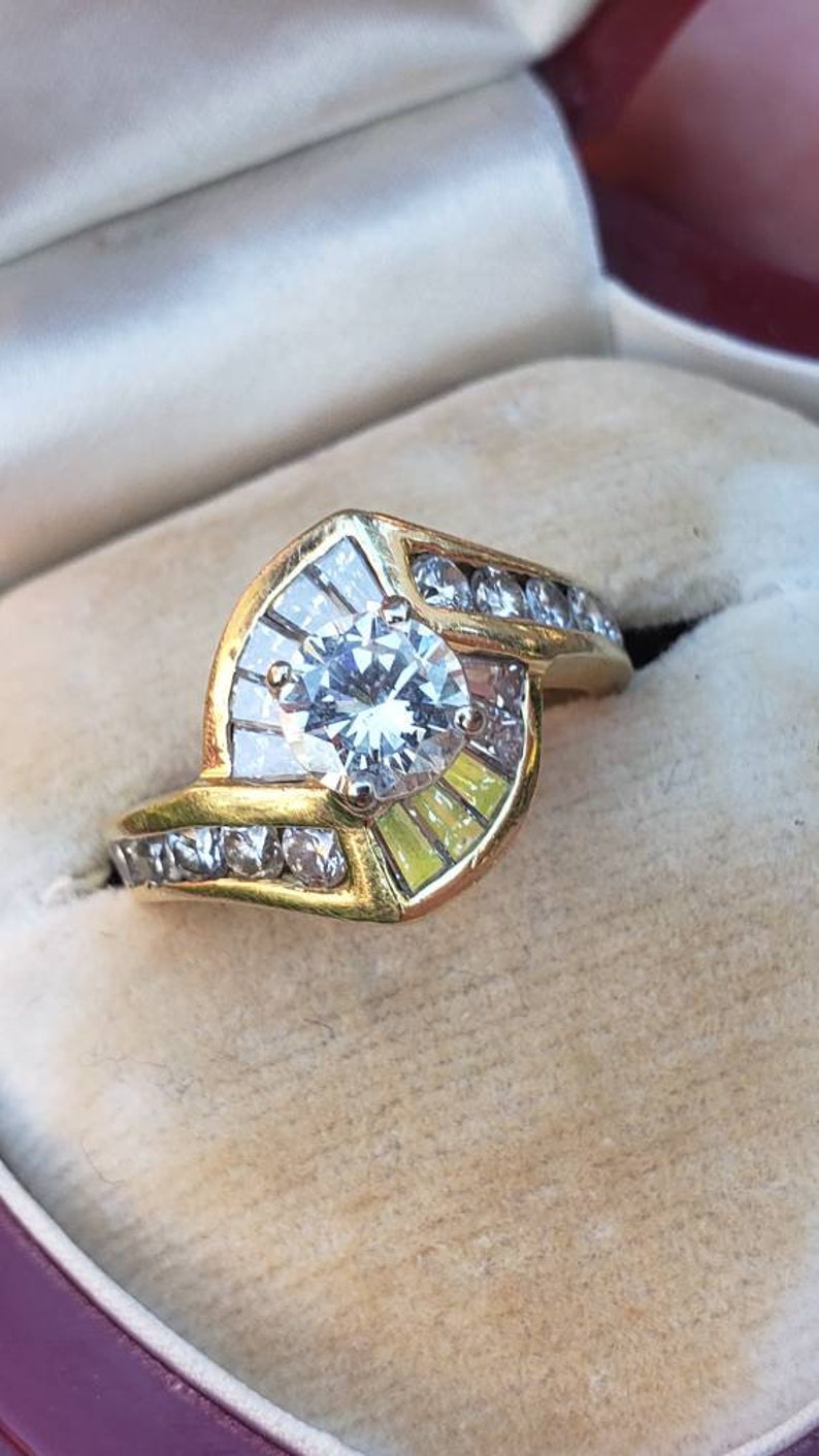 May include: A gold ring with a large, round, clear center stone surrounded by smaller clear stones. The ring is set in a curved, prong setting.