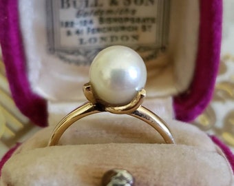 May include: A gold ring with a large, white pearl set in a simple, elegant design. The ring is displayed in a pink velvet box with a label that reads "Bull & Son, Goldsmiths, 182-184, Regent St, & 41, New Bond St, LONDON."