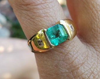 May include: A gold ring featuring a large, rectangular emerald gemstone. The ring has a classic design with small accent stones on either side of the emerald. The emerald is a vibrant green color.