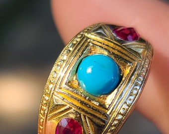 May include: A gold ring with a turquoise stone and two red stones. The ring has a detailed design with intricate carvings.