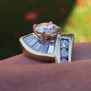 May include: A gold ring with a large round diamond in the center and smaller diamonds on the band. The ring is set in a fan shape with baguette diamonds.
