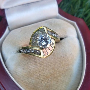 May include: A gold engagement ring with a large, round, clear diamond center stone. The ring has a unique design with smaller diamonds set in a curved band.