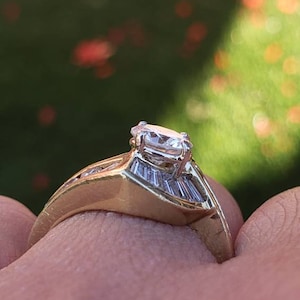 May include: A gold engagement ring with a large, clear, oval-shaped diamond in the center. The band is set with smaller diamonds on either side of the center stone.