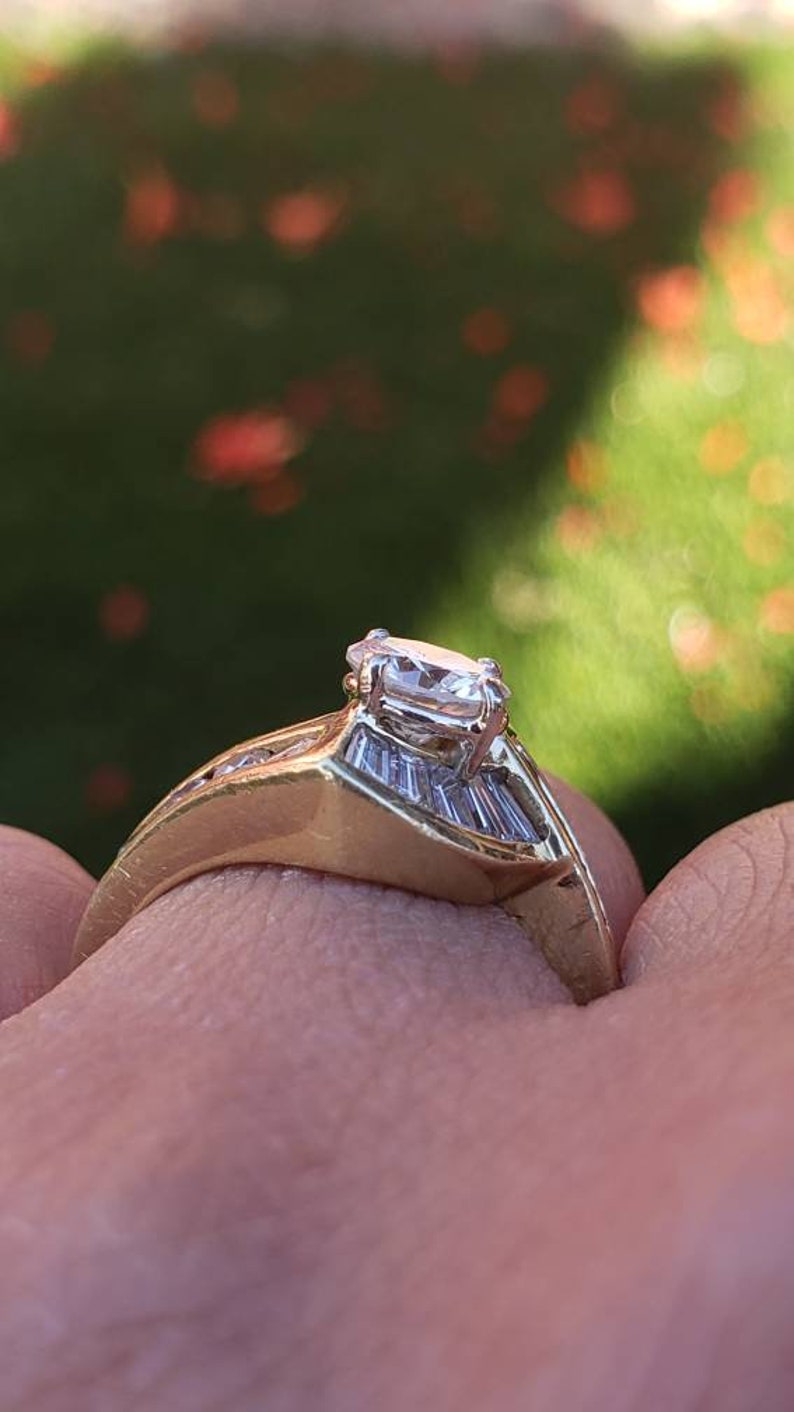 May include: A gold engagement ring with a large, clear, oval-shaped diamond in the center. The band is set with smaller diamonds on either side of the center stone.