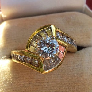 May include: A gold ring with a large, round, clear center stone surrounded by smaller, clear stones. The ring has a curved design and is set in a gold band.