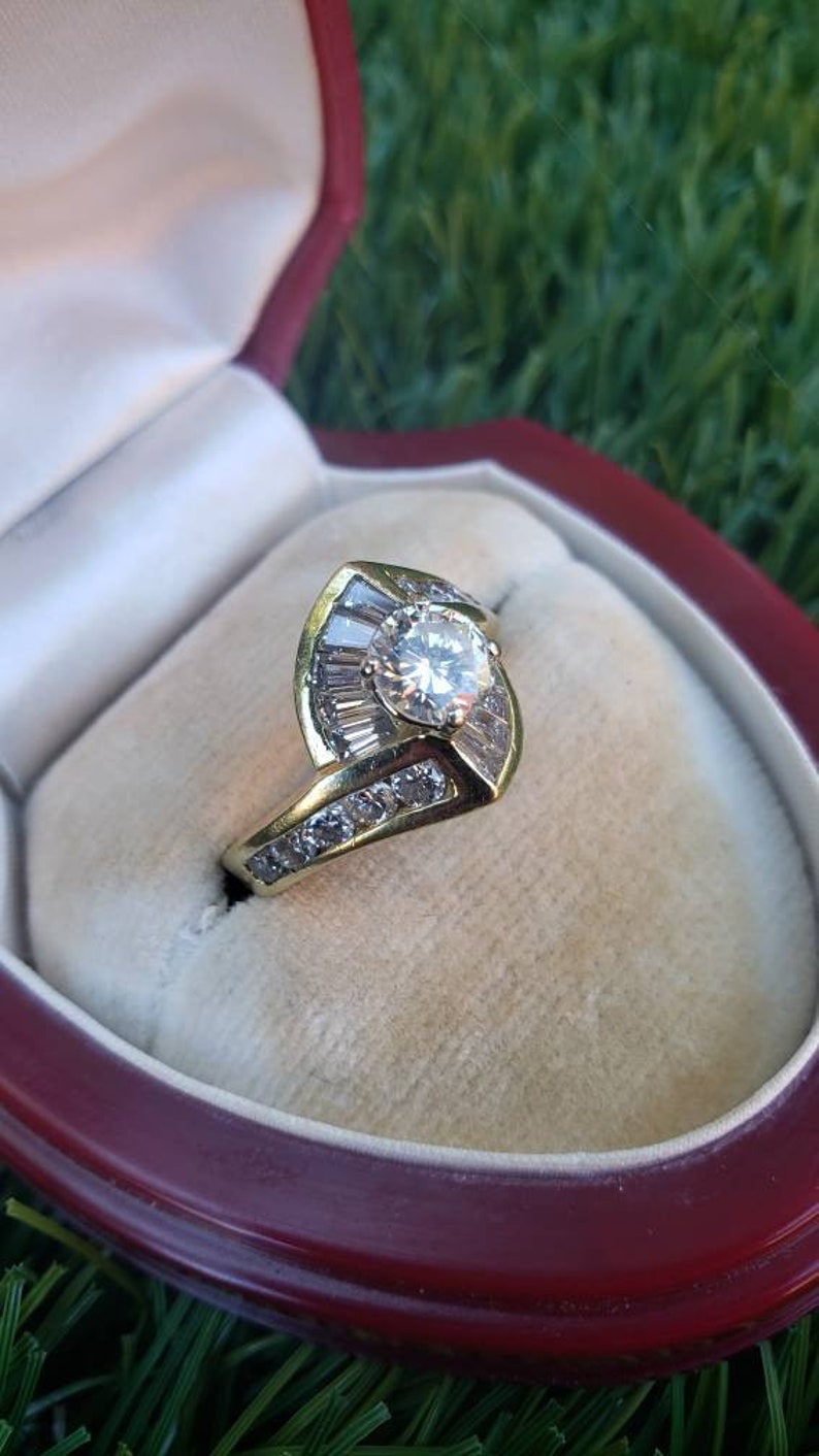 May include: A gold ring with a large round diamond surrounded by smaller diamonds in a leaf-shaped setting. The ring is in a red velvet ring box.