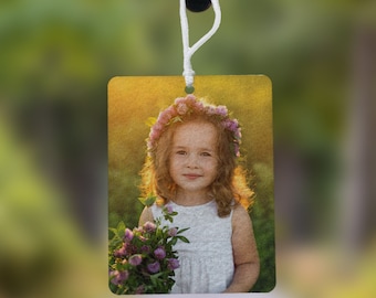 May include: A rectangular car air freshener hanging from a rearview mirror, featuring a printed photograph of a young child with curly hair wearing a flower crown and a white dress, while holding a bouquet of purple clover flowers in a field.