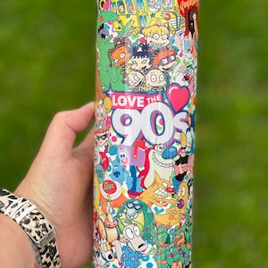 Könnte beinhalten: Ein weißer Edelstahlbecher mit einem bunten Design mit Cartoon-Charakteren aus beliebten Nickelodeon-Shows der 90er Jahre. Das Design enthält Figuren wie Ren & Stimpy, Rugrats, Doug und Rocko's Modern Life. Der Becher trägt den Text "LOVE THE 90s" mit einem roten Herzen.