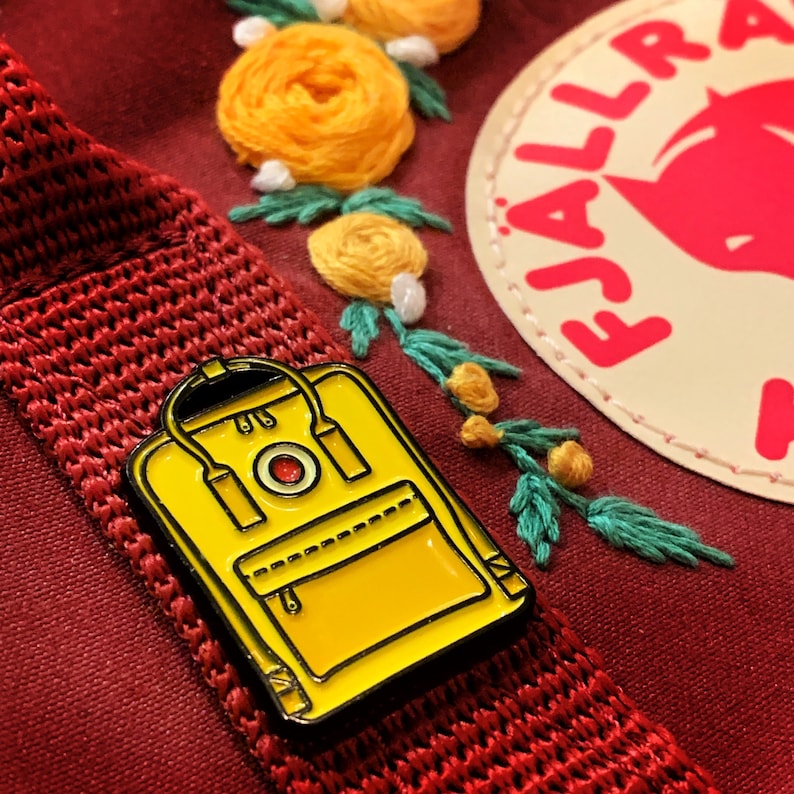May include: Yellow enamel pin of a classic backpack with a red circle logo. The pin is on a red fabric background with embroidered flowers.