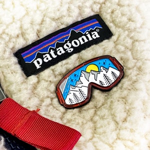 Może przedstawiać: Biała kurtka polarowa z naszywką z logo Patagonia w kolorze niebieskim i fioletowym oraz emaliowaną szpilką przedstawiającą pasmo górskie z zachodzącym słońcem.