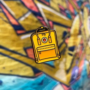 May include: Yellow enamel pin of a classic backpack with black straps and a red circle logo.
