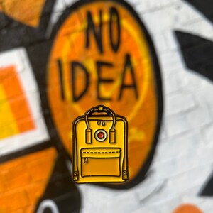 May include: A yellow enamel pin of a backpack with a red circle logo and the text "NO IDEA" in the background.