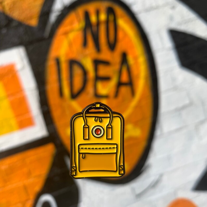 May include: A yellow enamel pin of a backpack with a red circle logo and the text "NO IDEA" in the background.
