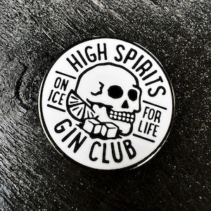 Puede incluir: Pin redondo en blanco y negro con el texto "HIGH SPIRITS FOR LIFE" y "GIN CLUB". El diseño incluye una calavera, una rodaja de limón y cubitos de hielo. Sobre una superficie de madera oscura.