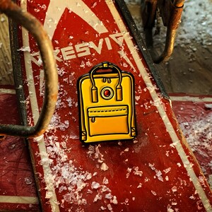 May include: A yellow enamel pin of a small backpack with a red circle and a white button on the front. The pin is on a red and white surface.