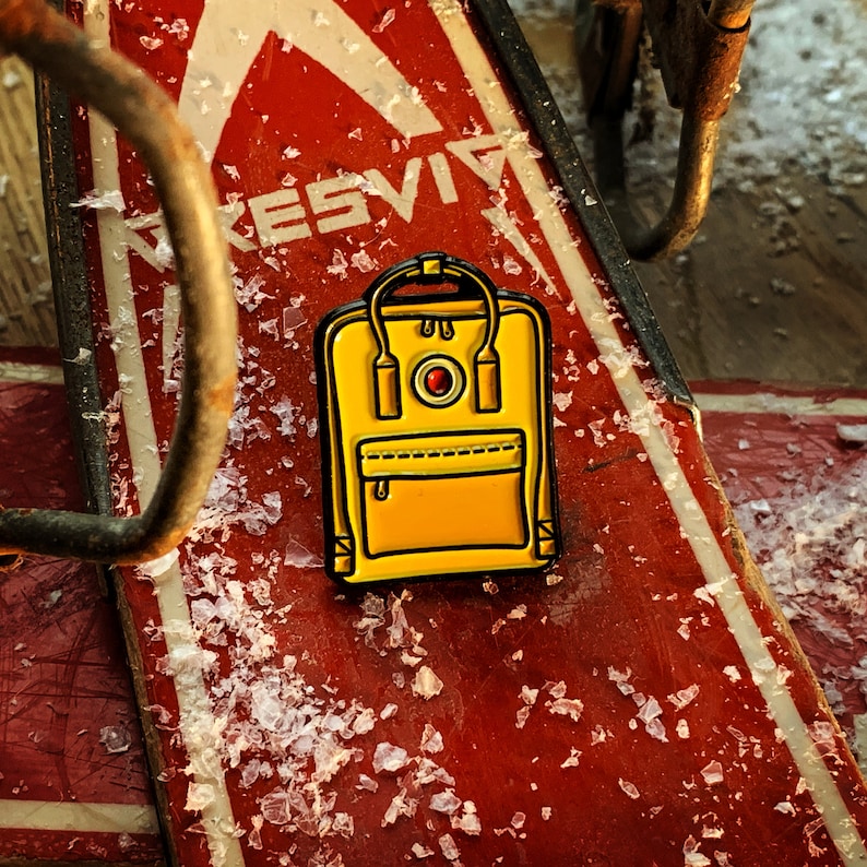 May include: A yellow enamel pin of a small backpack with a red circle and a white button on the front. The pin is on a red and white surface.