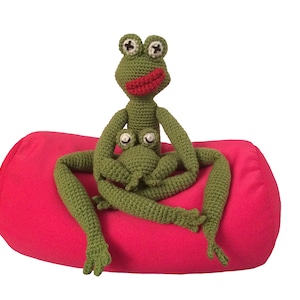 May include: Two green crocheted frog plushies sitting on a pink cylindrical pillow. The larger frog has a red mouth and the smaller frog is sitting on the larger frog's lap.