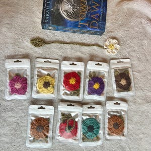 May include: A book titled "Tower of Dawn" with a crocheted bookmark and several crocheted flower ornaments in clear plastic packaging. The flowers are various colors, including pink, yellow, red, purple, and brown.