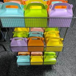 May include: A display of colorful gift boxes in various shades, including blue, green, orange, yellow, and purple. The boxes are arranged on a three-tiered metal wire rack, showcasing their vibrant hues and handle details. The boxes are ideal for party favors.