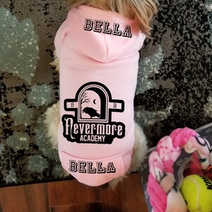 May include: A light pink pet hoodie featuring the name BELLA printed in black on the hood and lower back. The center back displays a black graphic of a raven inside an arched window with the text Nevermore ACADEMY, 19, and 91.