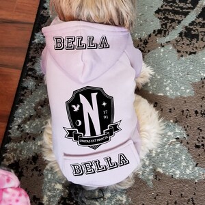 May include: A light purple pet hoodie featuring the name BELLA printed on the hood and lower back in a black collegiate font. A black shield emblem is centered on the back, displaying a large letter N, the year 1791, and the text UNITAS EST INVICTA.