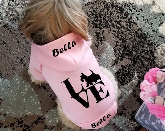 Puede incluir: Una sudadera con capucha de color rosa claro para perro con mangas largas. La parte posterior de la prenda muestra el texto negro Bella, una silueta negra de perro, la palabra LOVE con la silueta reemplazando la O y el nombre Bella abajo.