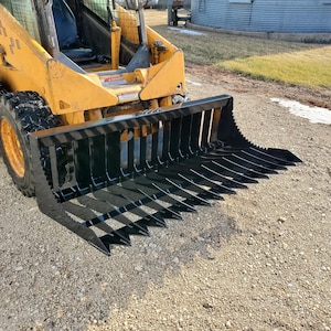 Puede incluir: Un accesorio de rastrillo para paisajismo de metal negro para una cargadora de dirección deslizante. El rastrillo tiene 12 dientes y se conecta a la cargadora mediante un sistema de enganche rápido.