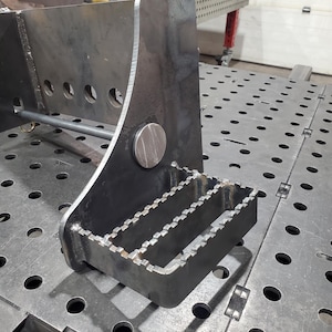 Pode incluir: Um close-up de um apoio para os pés de metal inacabado, provavelmente para máquinas ou uso industrial. O apoio para os pés tem uma forma retangular com uma superfície texturizada para aderência e é soldado a uma estrutura metálica maior. O metal tem uma aparência industrial bruta.