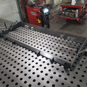 May include: A metal welding table with a grid pattern and a metal frame being assembled on top. The frame is held in place with clamps.