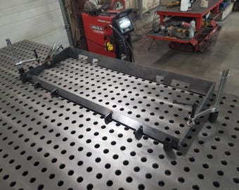 May include: A metal welding table with a grid pattern and a metal frame being assembled on top. The frame is held in place with clamps.