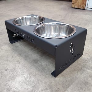 Puede incluir: Un soporte negro de metal para cuencos de mascotas con dos cuencos de acero inoxidable. El soporte tiene texto recortado que dice "DUNCAN" y "SHOP DOG". El soporte está diseñado para elevar los cuencos de comida y agua.