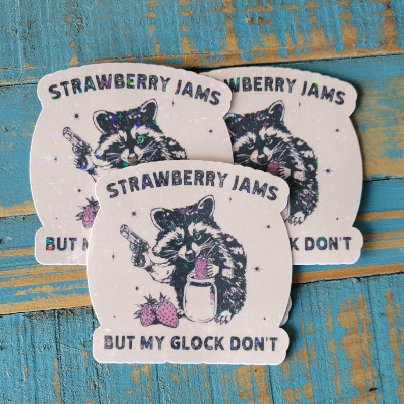 May include: Three stickers with a raccoon graphic holding a gun and a jar of jam. The text on the stickers reads "STRAWBERRY JAMS BUT MY GLOCK DON'T". The stickers have a holographic finish and a light background.