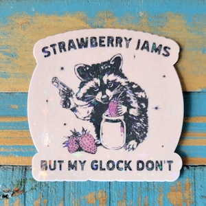 May include: A sticker with a raccoon illustration holding a gun and a jar of strawberries. The text reads "STRAWBERRY JAMS BUT MY GLOCK DON'T". The sticker has a holographic effect and a white background.
