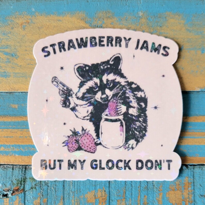 May include: A sticker with a raccoon illustration holding a gun and a jar of strawberries. The text reads "STRAWBERRY JAMS BUT MY GLOCK DON'T". The sticker has a holographic effect and a white background.