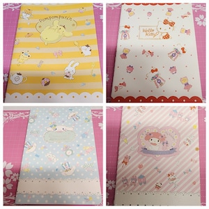 May include: Four different colorful notebooks with various cartoon characters. The notebooks feature Pompompurin, Hello Kitty, and Cinnamoroll. The notebooks have a yellow, white, blue, and pink background.