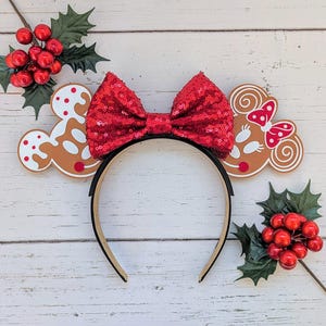 Op de afbeelding: Minnie Mouse-oorkleppenband met een gingerbread-thema en een rode pailletten strik. Het hoofdband is bruin en wit met een gingerbread man-ontwerp. De strik is rood en glinstert.