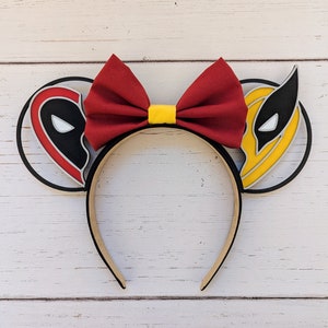 May include: A pair of black mouse ears with a red bow and yellow center. One ear features a black and white Deadpool character and the other ear features a yellow and black Wolverine character.