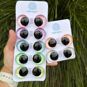 May include: A selection of colourful glass eyes for crafts, in various sizes. The eyes are in shades of pink, purple, blue, green, and yellow-green, with black pupils. Each card is labelled with the text "YARN WITH" and size measurements in millimetres.